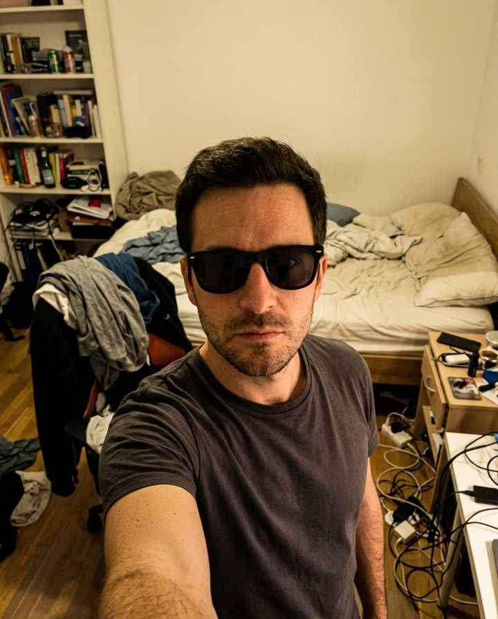 Man taking a selfie with sunglasses in a dim, messy bedroom